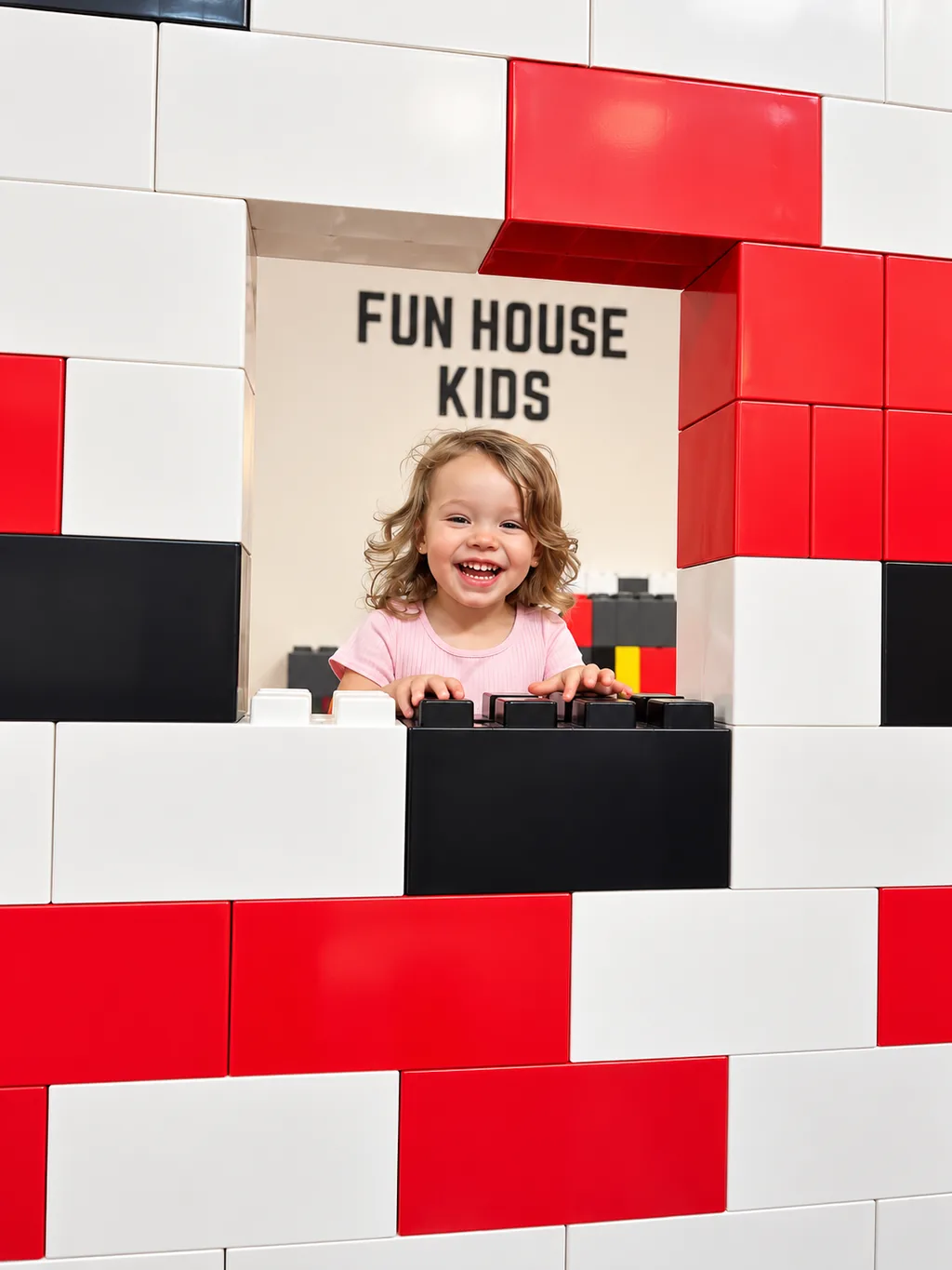 Smiling child peeking through a window in a wall of large red, white, and black blocks.