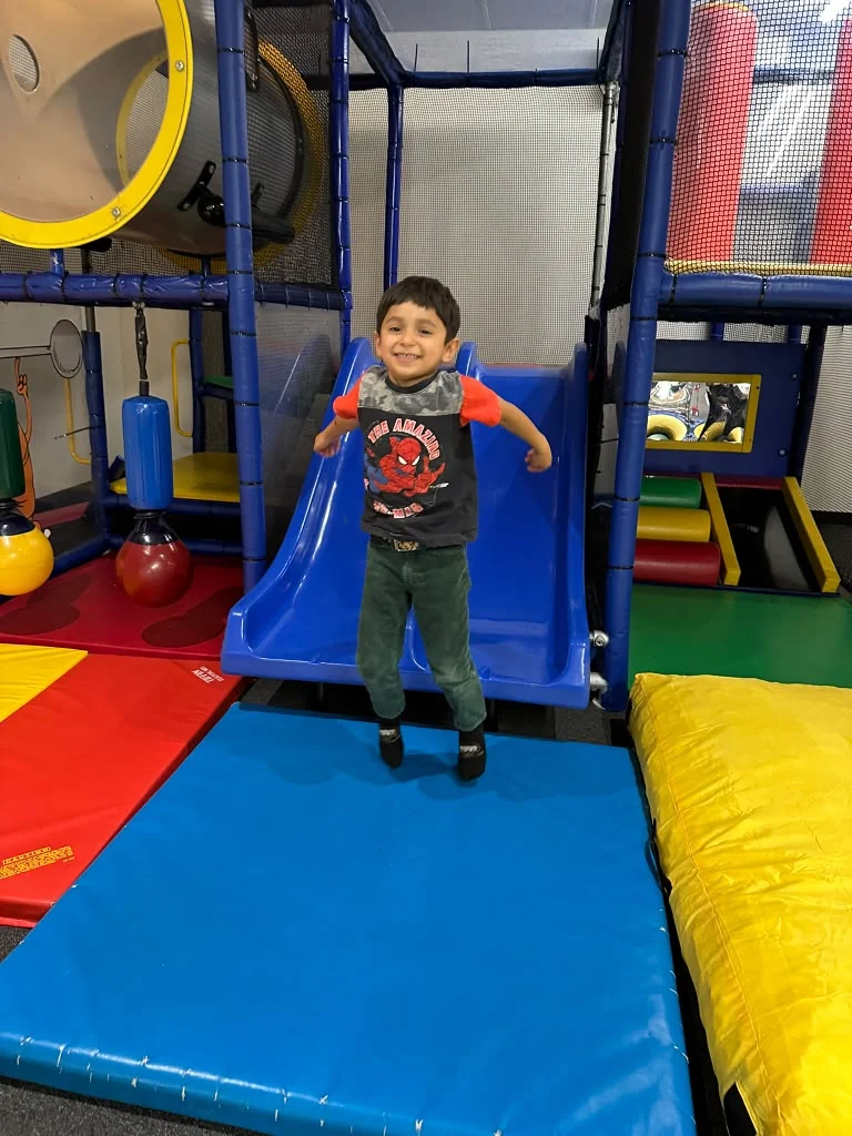 Child at the bottom of the blue slide beside colorful padded mats.