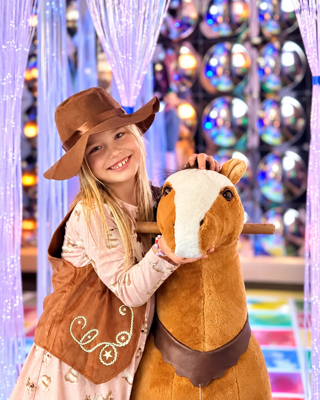 Smiling child in a cowboy hat and vest hugging a toy horse in the glowing light room.