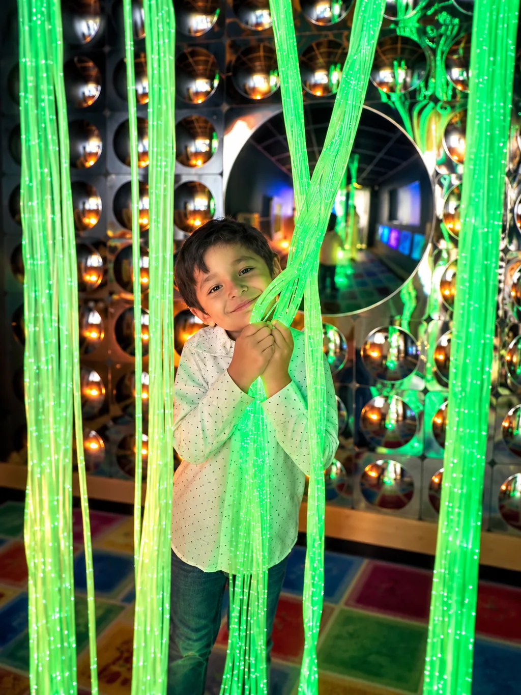 Child among glowing green light strands in front of a mirrored wall.
