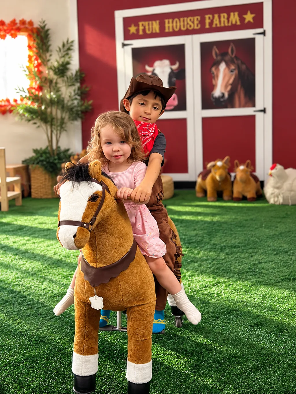 Two children in cowboy hats riding a toy horse in front of the Fun House Farm barn.