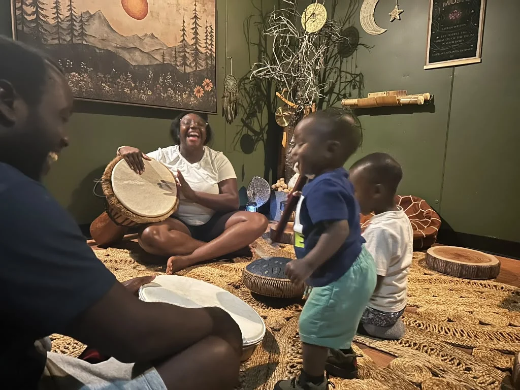 Children and an adult playing hand drums together in a decorated indoor space.