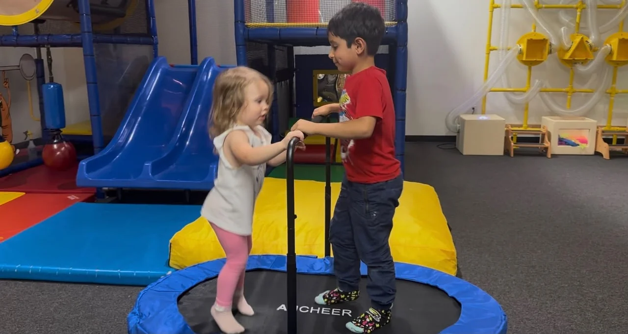 Two children standing on a small trampoline beside a blue slide.